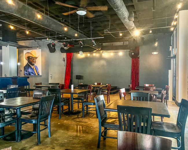 The image shows an interior view of a restaurant with wooden tables and chairs, featuring a large portrait on the wall and overhead lights illuminating the space.
