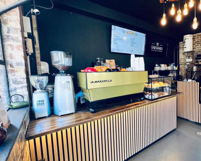The image shows a modern coffee shop interior with a service counter featuring a green espresso machine, display cases of pastries, and a menu board on the wall.