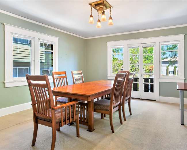 The image shows a spacious dining room featuring a wooden table surrounded by six chairs, with large windows allowing natural light and a side table with a small appliance.