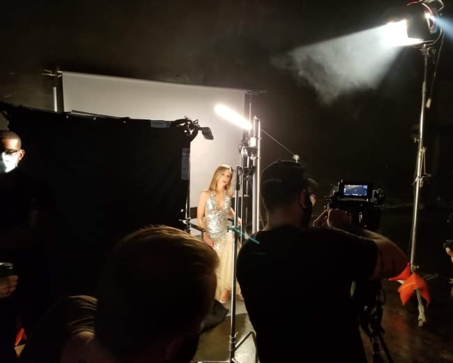 A woman in a sparkling dress stands in a spotlight on a film set, while crew members operate cameras and equipment around her.