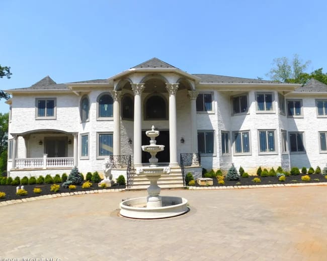 A large, multi-story mansion features a central entrance with four columns and a circular fountain in the driveway.