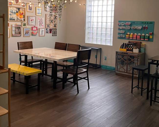 The image shows a brightly lit interior space with two tables, a gallery wall of framed pictures, and a variety of seating arrangements.