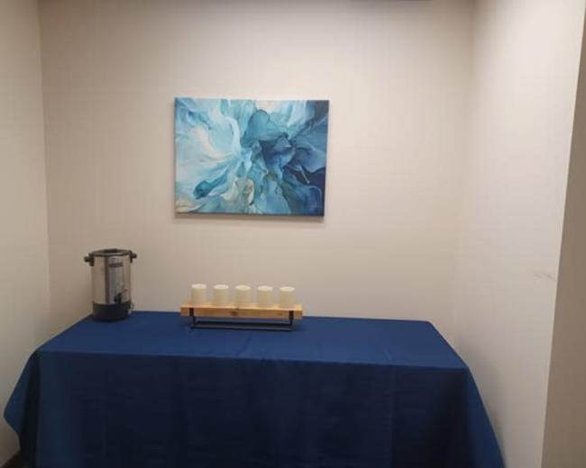 A blue tablecloth covers a table holding four candles and a water dispenser, with a blue abstract painting on the wall above.