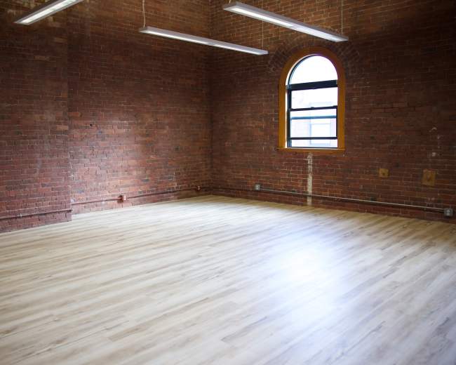 An empty room with exposed brick walls and a large window letting in natural light, featuring a wooden floor.