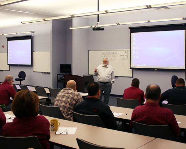 A speaker stands in front of an audience in a classroom setting, with projector screens displaying a presentation.