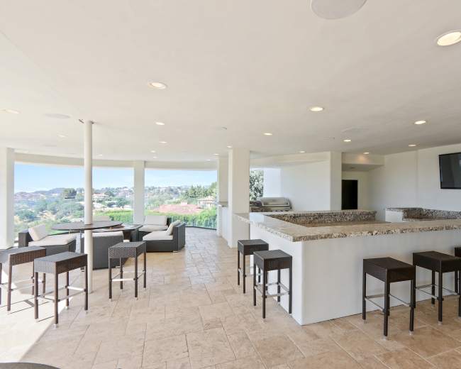 The image shows a modern outdoor bar area with a granite countertop, seating for eight, and a scenic view beyond.