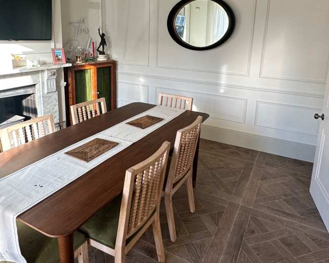 A wooden dining table with six chairs is set in a room featuring green walls, a round mirror, and a woven light fixture.