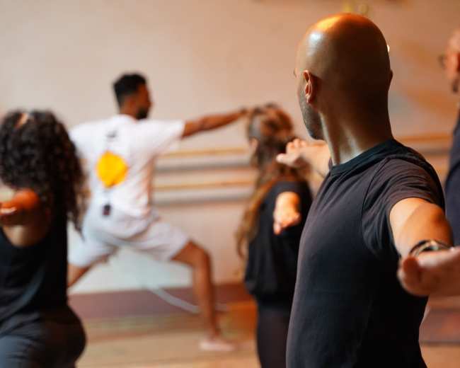 A group of individuals participates in a dance class, with some facing the camera and others performing movements in the background.