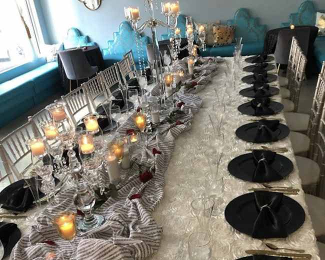 A long dining table is set with black plates, glassware, and candles, adorned with a striped cloth and decorative centerpieces.