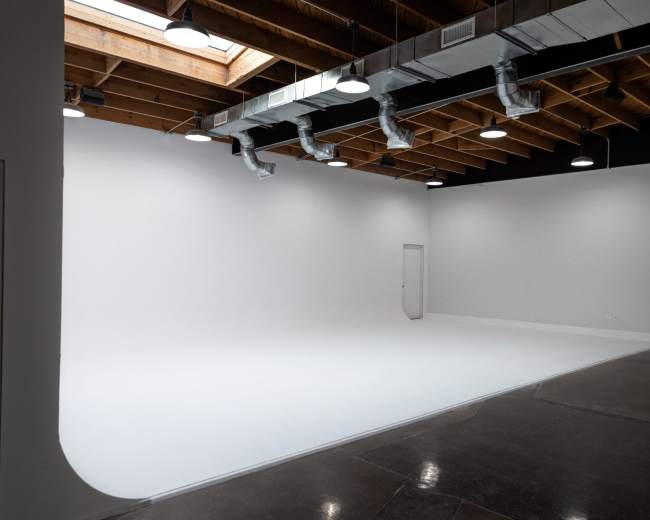 The image shows a spacious, empty studio with a white backdrop, wooden ceiling beams, and overhead lighting.