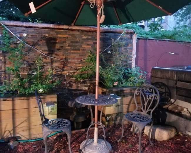 A small round table with two chairs is placed under a green umbrella beside a stone wall and garden planters.