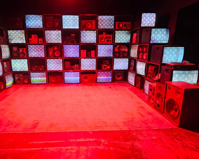 The image shows a room filled with stacked speakers and monitors arranged in a grid pattern, illuminated by red lighting.