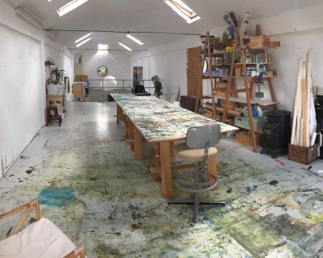 The image shows a spacious art studio with a large table covered in paint splatters, shelves filled with art supplies, and various works in progress along the walls.