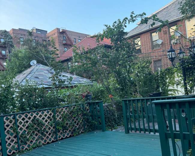 The image shows a green wooden deck surrounded by lush greenery and urban buildings in the background.