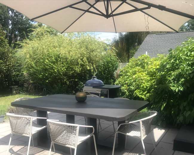 A patio table with four white chairs under a large umbrella, surrounded by greenery and a barbecue grill nearby.