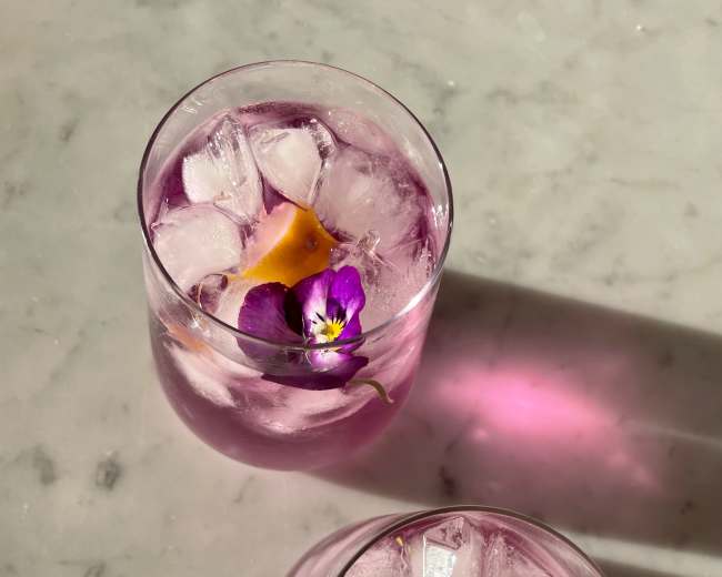 Two glasses of purple-hued drinks with ice, garnished with lemon and a pansy flower, sit on a marble surface.