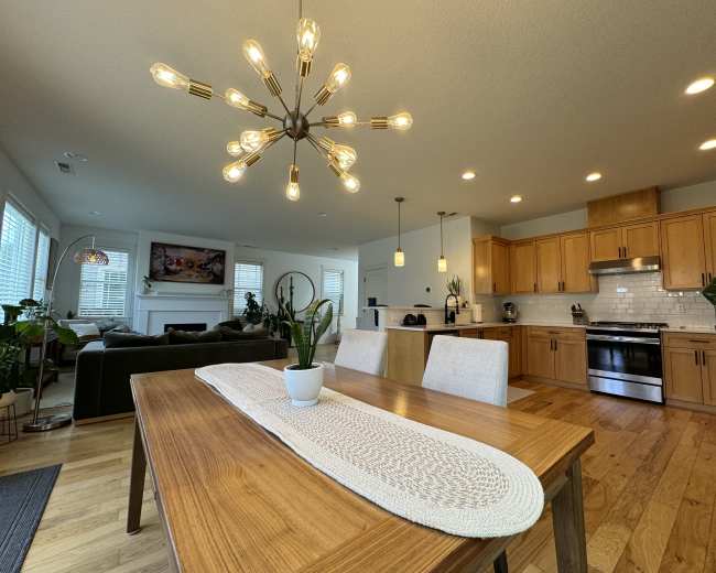A modern dining area features a wooden table with a centerpiece plant, surrounded by two chairs, and is adjacent to a kitchen and a cozy living room.