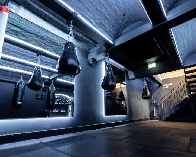 The image shows a dimly lit gym interior featuring several hanging punching bags, mirrored walls, and a staircase.