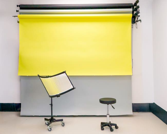 The image shows a photography studio with a bright yellow backdrop and a gray one, along with a black stool and a reflective screen on a wheeled stand.