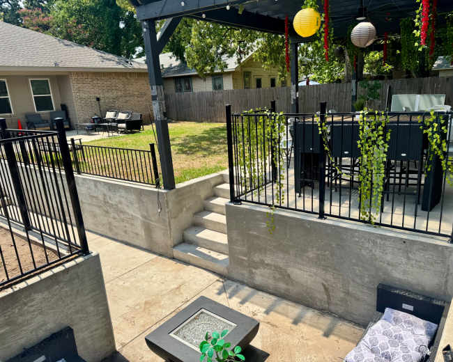 The image shows a landscaped outdoor space with a concrete patio, steps leading to a grassy area, and a covered seating area with furniture.