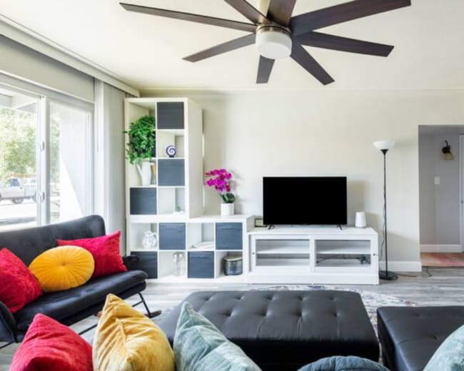 The living room features a modern design with a black ceiling fan, a dark sofa, colorful cushions, a sleek entertainment unit, and large windows allowing natural light.