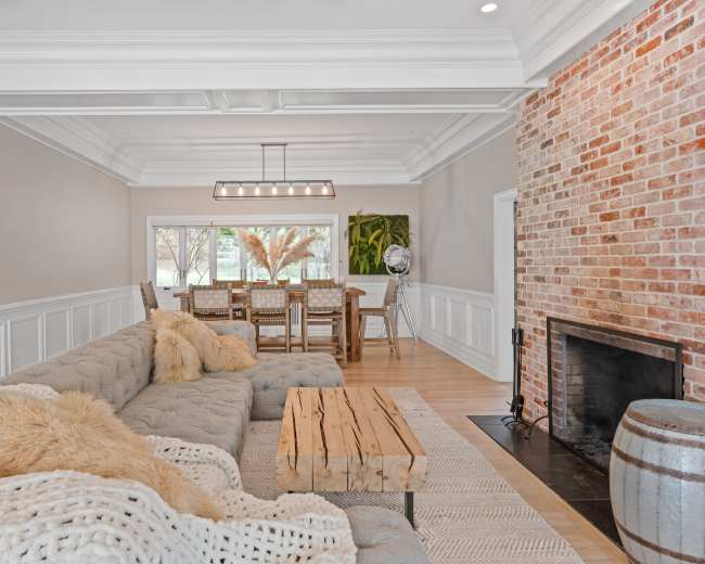 A spacious living room features a large sectional sofa, a wooden coffee table, and a brick fireplace, with a dining area visible through a large window.