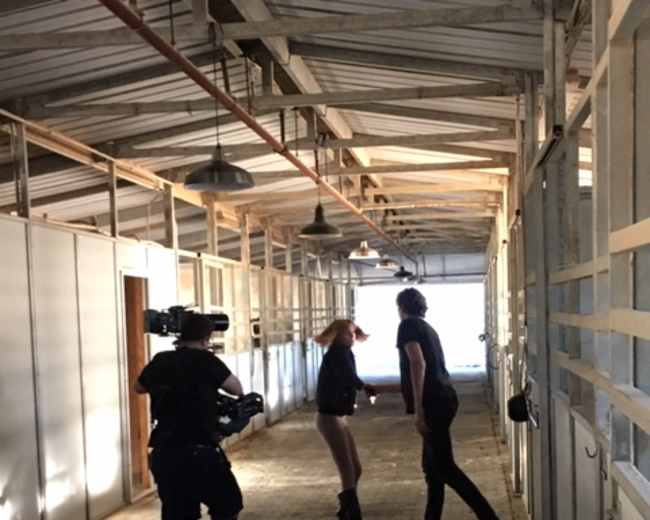 A camera operator records two people interacting in a well-lit corridor lined with horse stalls.