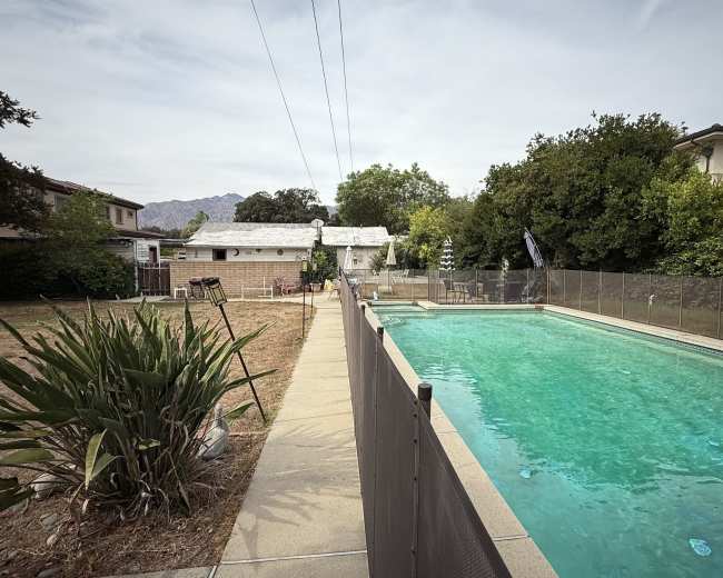 A swimming pool bordered by a chain-link fence is situated alongside a grassy area and residential buildings.