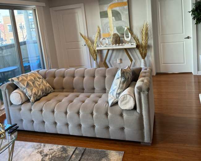 The image shows a modern living room with a light gray tufted sofa, decorative pillows, a console table with a decorative arrangement, and large windows allowing natural light.