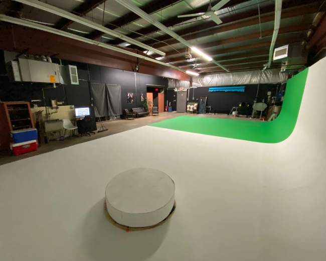 A spacious filming studio with a white floor, a green screen backdrop, and various equipment scattered around.