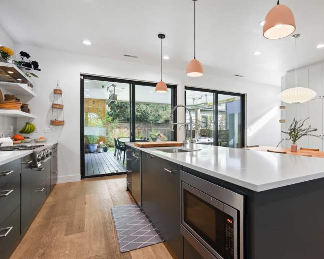 A modern kitchen features an island with a sink and microwave, light fixtures above, and large glass doors opening to an outdoor space.
