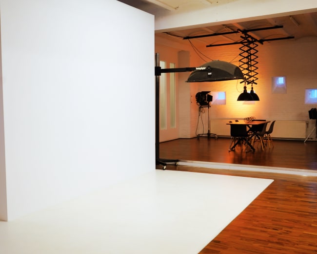 The image shows a photography studio featuring a white backdrop, a large light fixture, and a table with chairs in a well-lit space.