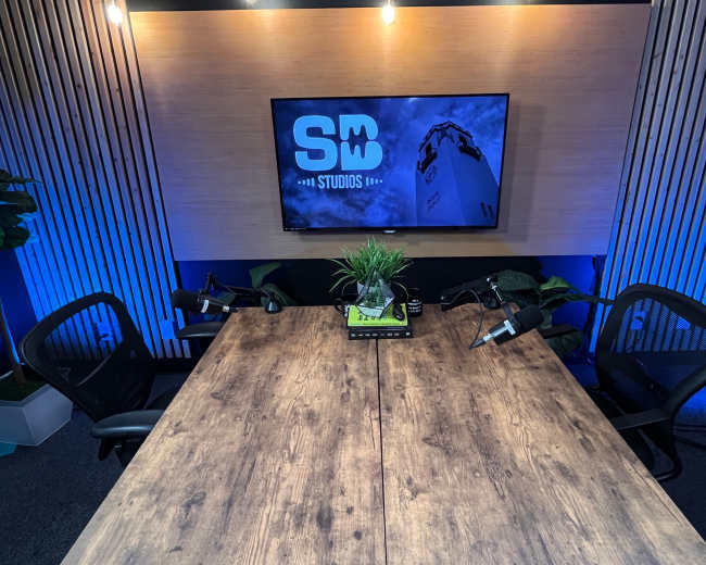 A wooden table is set up in a studio with two microphones and a television displaying the logo "SB STUDIOS" on the wall behind it.