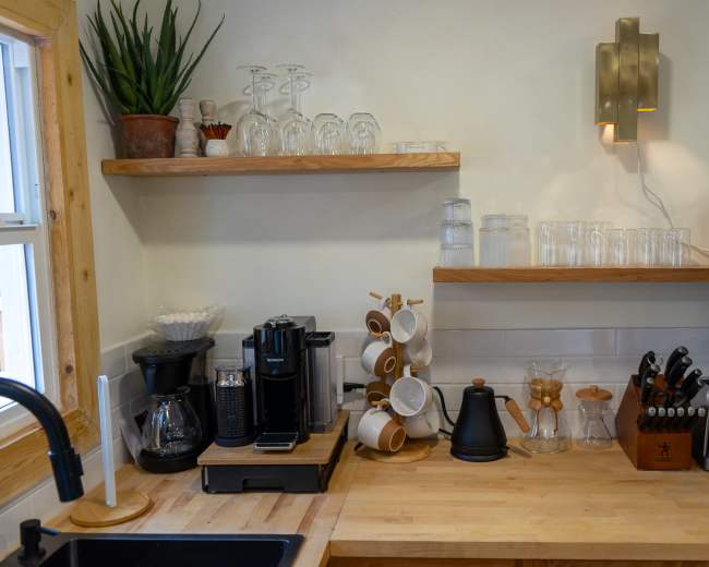 The image shows a modern kitchen countertop featuring a coffee maker, a kettle, various glassware, and a toaster, with wooden shelves above displaying more kitchen items.