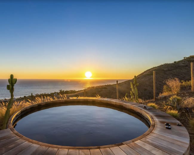 A circular wooden hot tub overlooks the ocean at sunset, framed by desert vegetation.