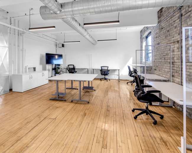 The image shows a modern office space with hardwood floors, featuring several desks and chairs, a large screen, and glass partitions.
