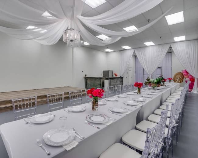 A spacious event room featuring a long dining table set with white tablecloths and floral arrangements, flanked by transparent chairs, with draped fabric ceiling decor and a small stage at one end.