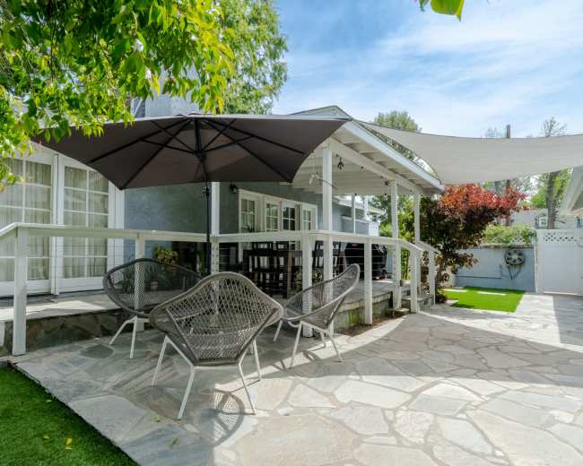 A patio features four wicker chairs and a large umbrella, surrounded by green grass and plants.