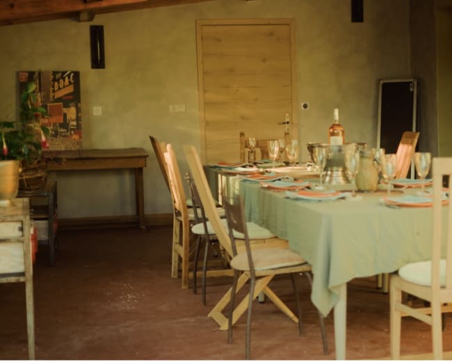 A long dining table is set in a softly lit room, with chairs arranged around it, and various dinnerware and glasses placed on the table.