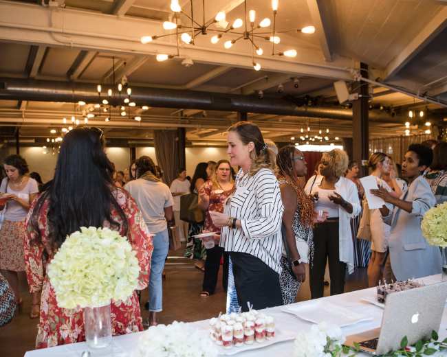 A group of people mingles at a well-lit event space featuring decorated tables with flowers and refreshments.