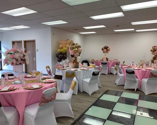 The image shows a decorated event space with circular tables set with pink tablecloths, white chairs adorned with gold sashes, and balloon arrangements in the corners.