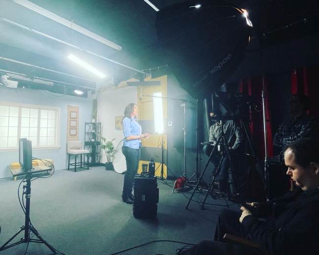 A woman stands in a well-lit studio, presenting while cameras and lights are set up around her.