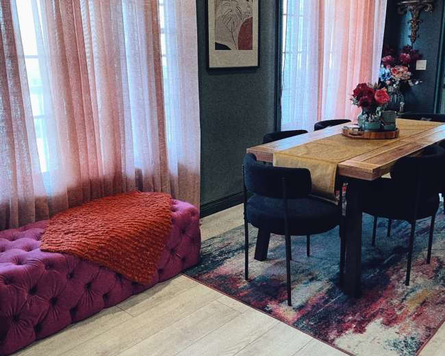 The image shows a modern dining area with a wooden table surrounded by black chairs, a pink tufted bench, and large windows dressed with sheer pink curtains.