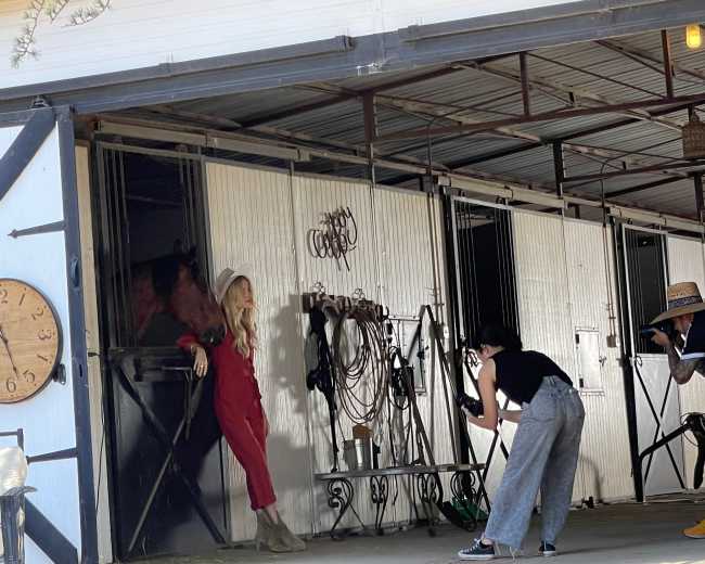 A model poses in a red outfit inside a barn while a photographer captures the scene and another person adjusts equipment.