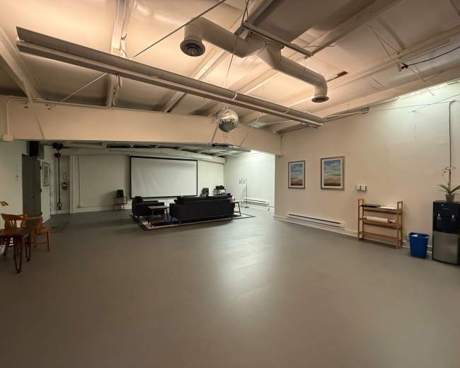The image shows a spacious, minimalist indoor area featuring a sofa set, a projector screen, two framed pictures on the wall, and some furniture arranged along the sides.