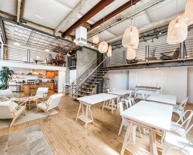 The image shows a spacious, modern loft with an open layout featuring wooden floors, a kitchen area, dining tables, and decorative pendant lights.