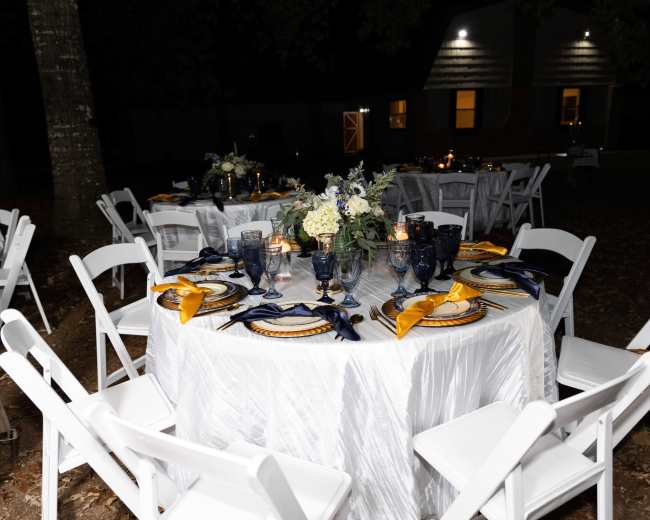 A beautifully set dining table with white chairs is arranged outdoors, featuring elegant blue and gold tableware amid floral centerpieces and soft lighting.