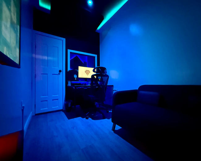 The image shows a dimly lit room featuring a desk with a computer setup, an office chair, and a couch against one wall.