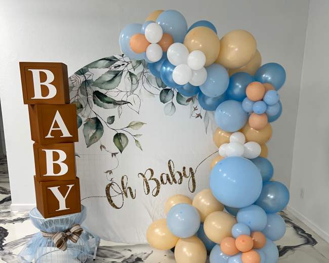 A decorative setup for a baby shower, including a large sign that reads "Oh Baby" and an arch made of blue, white, and beige balloons.