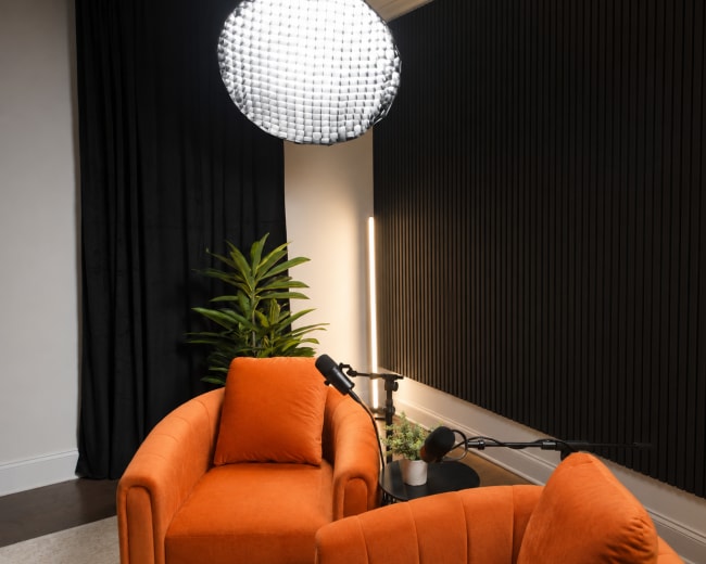 The image shows a modern podcast studio featuring two orange armchairs, a large pendant light, and a microphone setup against a dark, textured wall.